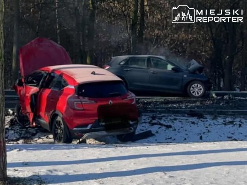 Czerwony samochód z uszkodzeniami stoi na poboczu drogi, a obok widać dymiący, niebieski pojazd w lesie.