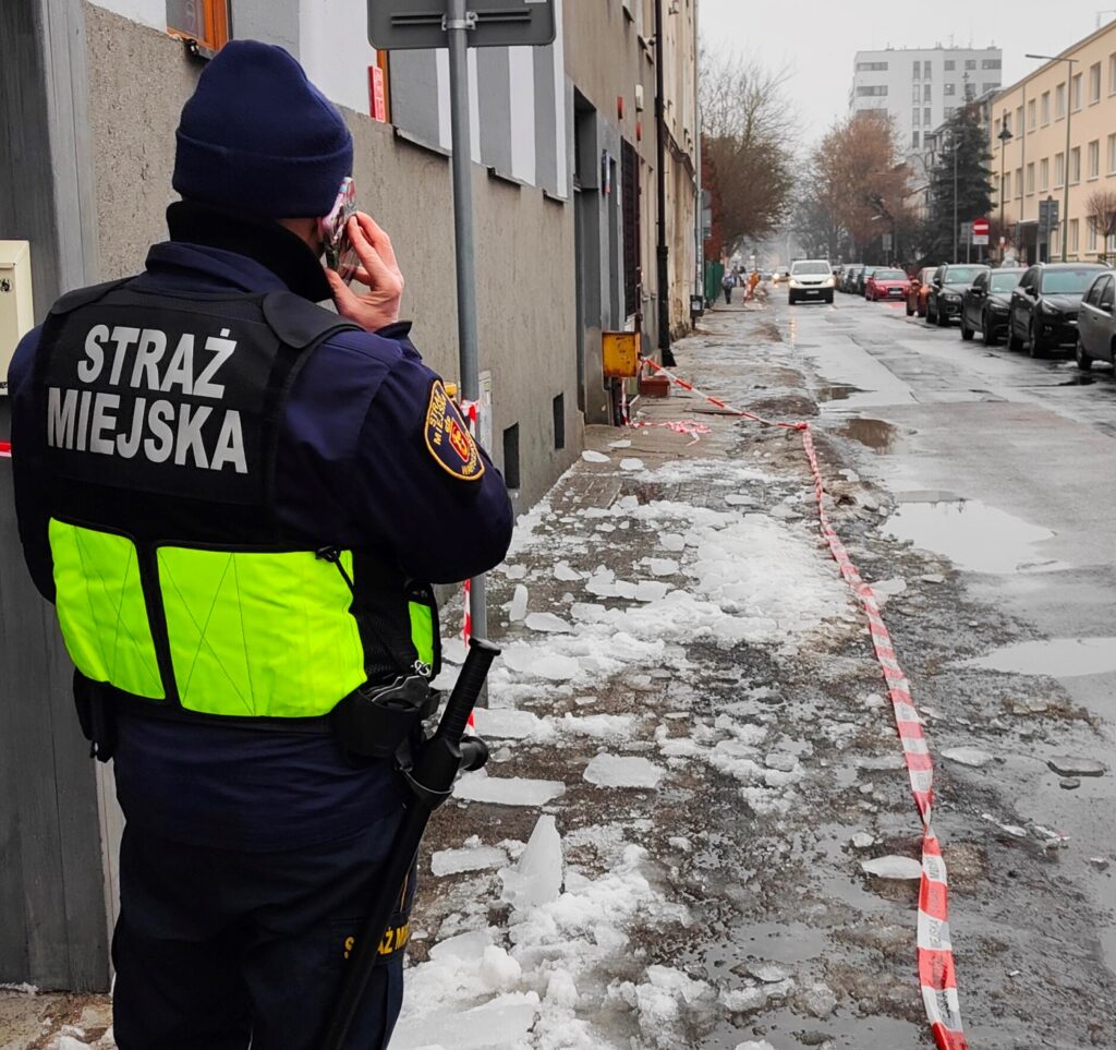 Funkcjonariusz straży miejskiej rozmawia przez telefon, stojąc obok zamarzniętej powierzchni na ulicy, otoczonej taśmą ostrzegawczą.
