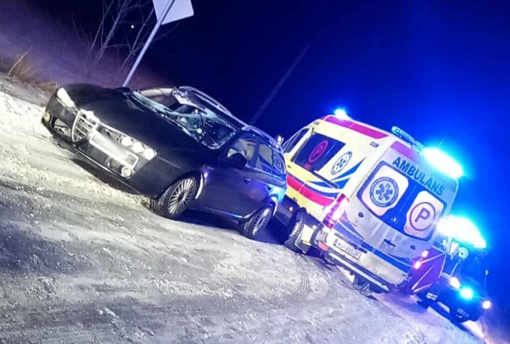 Czarny samochód osobowy zatrzymany na zaśnieżonej drodze obok karetki pogotowia z włączonymi sygnałami świetlnymi.