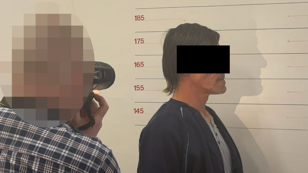 Mężczyzna w ciemnym ubraniu stoi na tle ściany z miarką, podczas gdy fotograf wykonuje mu zdjęcie.