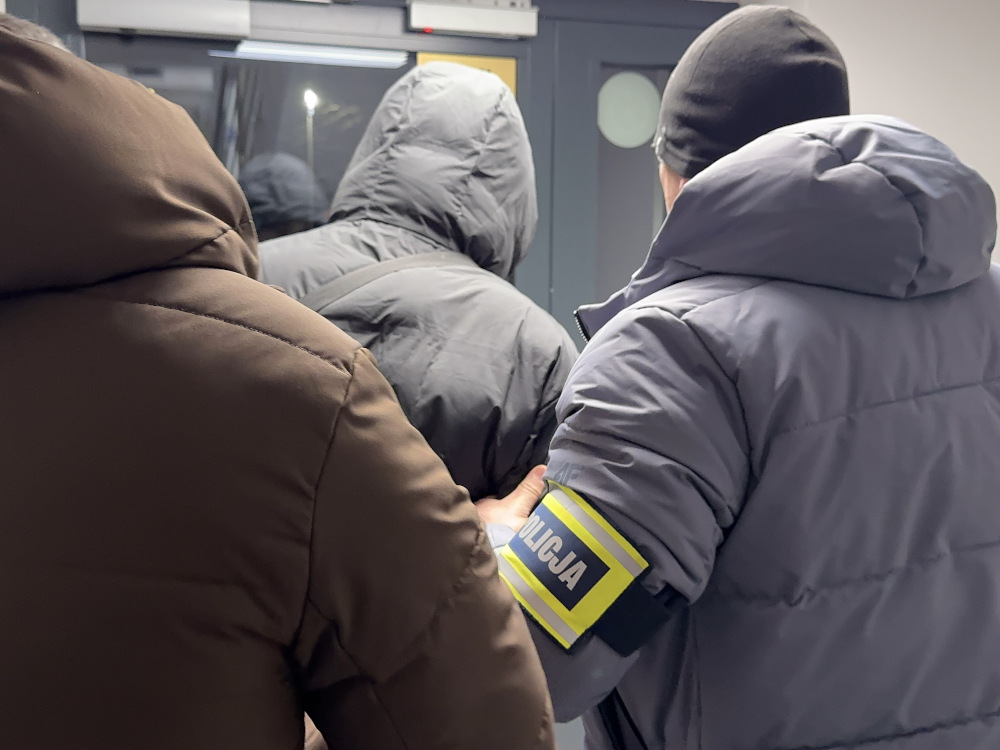 Mężczyzna w szarej kurtce jest prowadzony przez funkcjonariusza policji w mundurze, w zamkniętym pomieszczeniu.