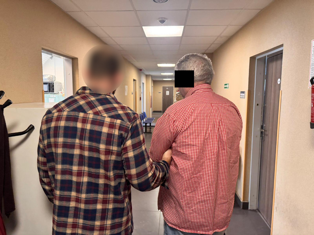 Mężczyzna w koszuli w kratę prowadzi drugiego mężczyznę w czerwonej koszuli przez korytarz w budynku.