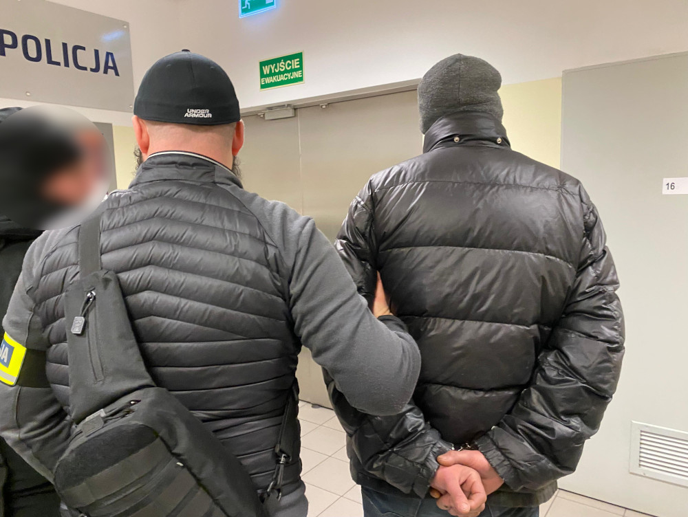 Mężczyzna w czarnej kurtce puchowej jest prowadzony przez funkcjonariusza policji w budynku administracyjnym.