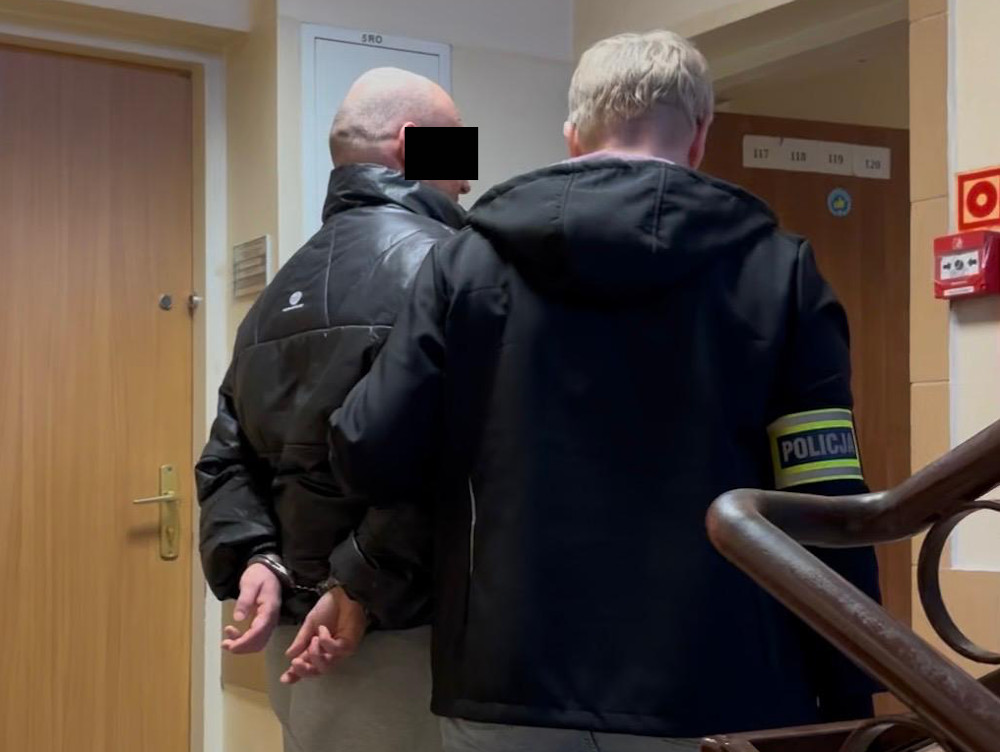 Mężczyzna w kajdankach prowadzony przez policjanta w korytarzu budynku mieszkalnego.