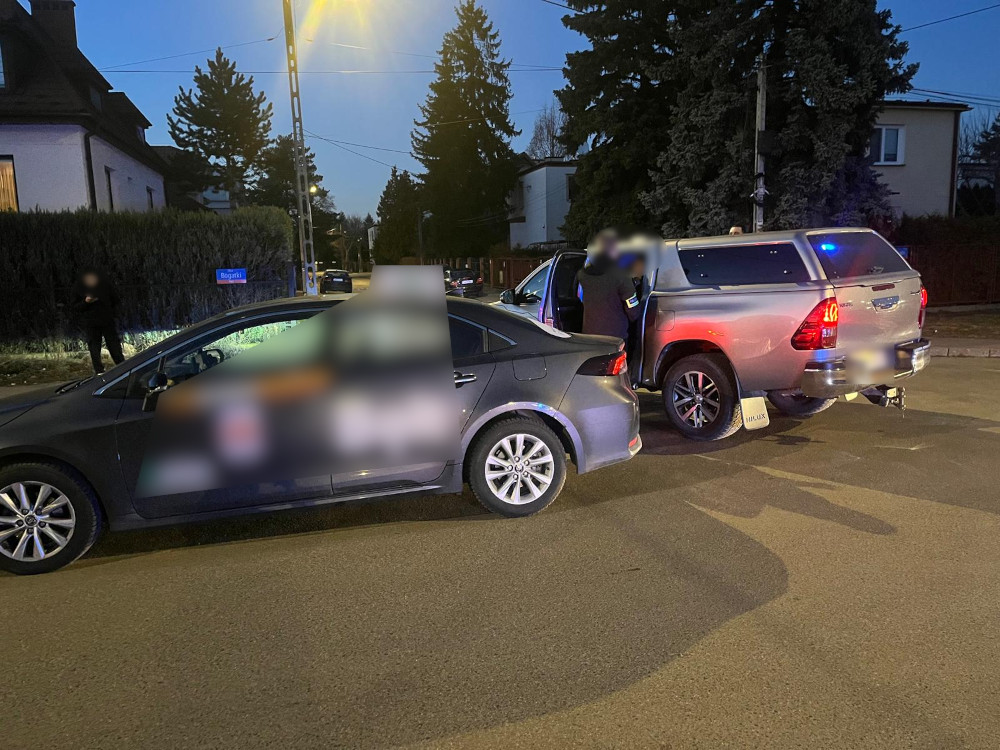 Mężczyzna w ciemnym ubraniu stoi obok zaparkowanego samochodu osobowego na ulicy, obok drugiego pojazdu z włączonymi światłami.