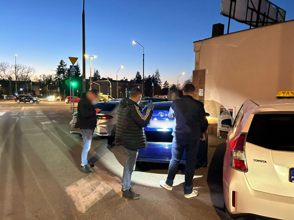 Grupa trzech mężczyzn rozmawia przy zaparkowanym samochodzie osobowym na skrzyżowaniu ulic w czasie zmierzchu.