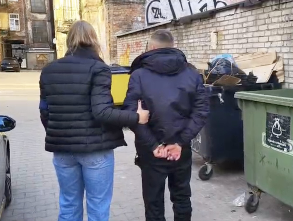 Kobieta w czarnej kurtce prowadzi mężczyznę z rękami związanymi z tyłu w wąskiej uliczce, obok kontenerów na śmieci.