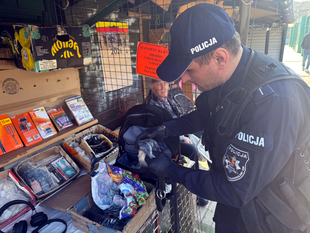 Policjant w mundurze przeszukuje torbę na stoisku z różnymi przedmiotami, a w tle siedzi starsza osoba.