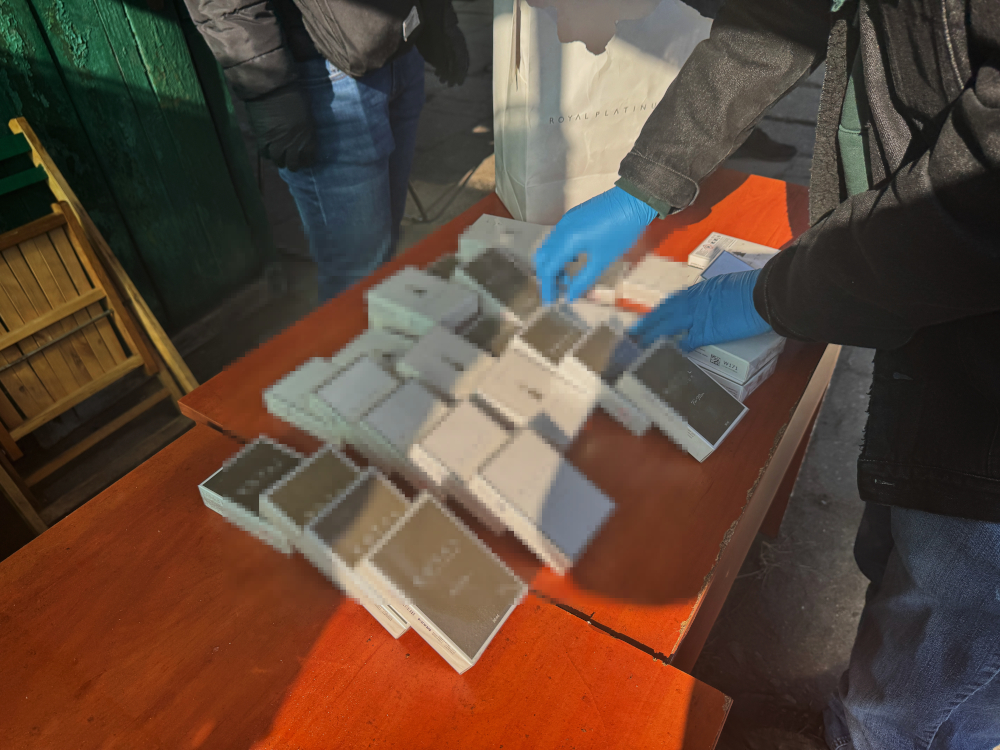 Osoba w rękawiczkach niebieskich układa pudełka na drewnianym stole w otoczeniu zielonej ściany.