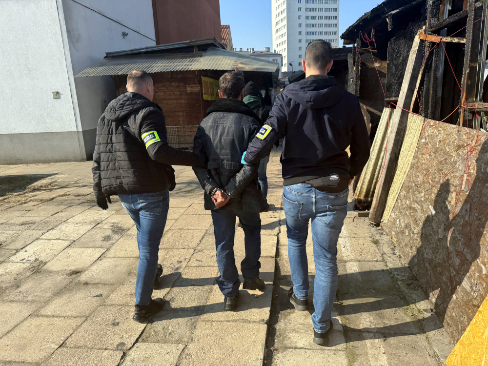 Trzej funkcjonariusze prowadzą mężczyznę z rękami związanymi z tyłu przez teren z drewnianymi budynkami.