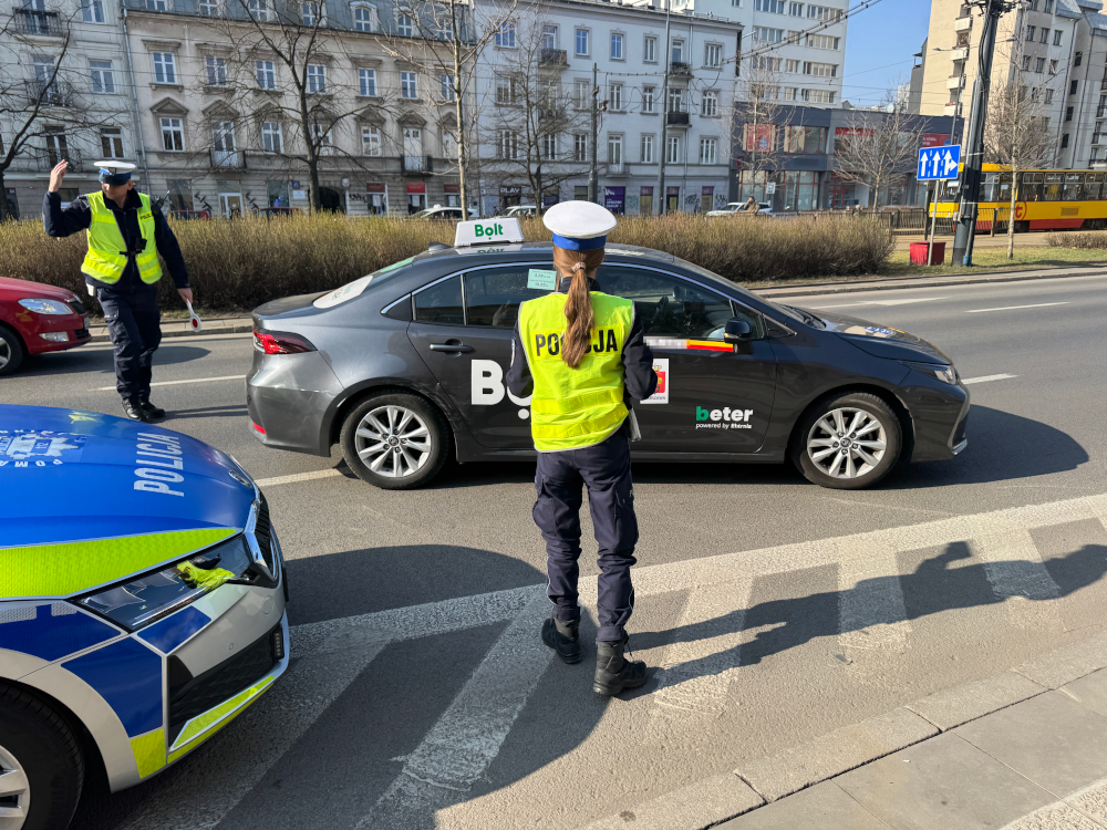 Policjant w kamizelce odblaskowej kieruje ruchem drogowym obok zatrzymanego samochodu z napisem "Bolt".