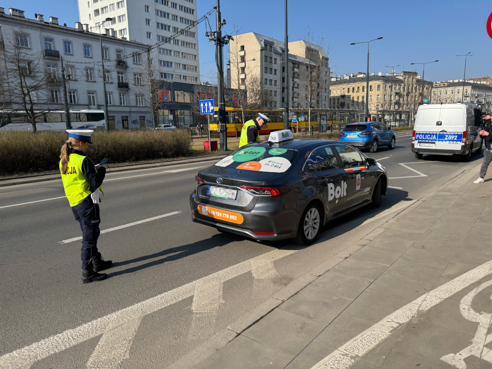 Policjantka w kamizelce odblaskowej sprawdza dokumenty kierowcy taksówki Bolt na ruchliwej ulicy.