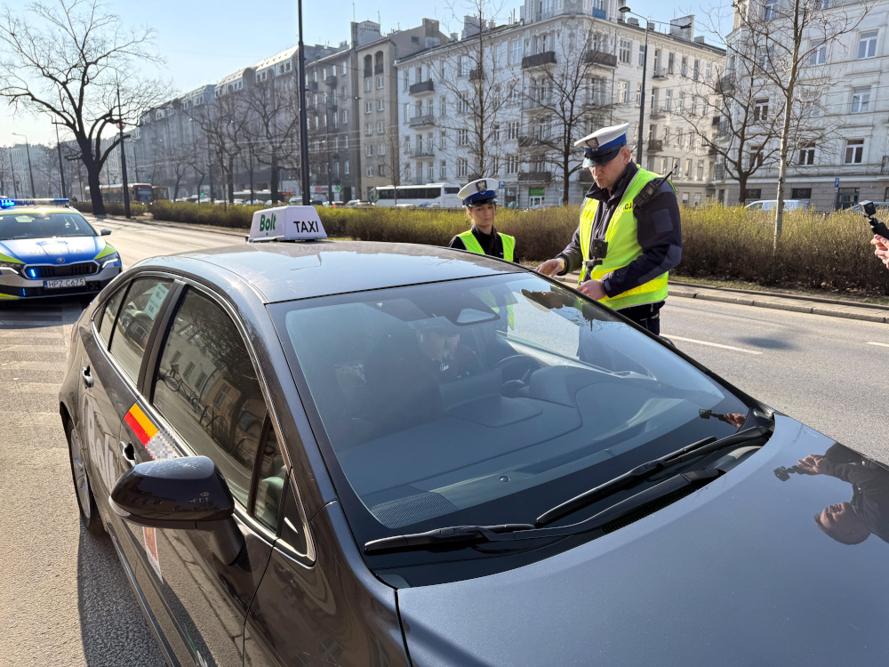 Policjanci w kamizelkach odblaskowych kontrolują taksówkę z logo Bolt na ruchliwej ulicy.
