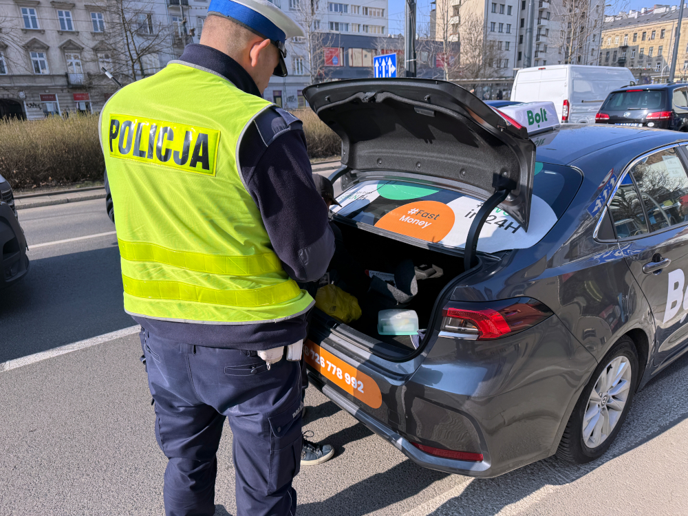 Policjant w kamizelce odblaskowej przeszukuje bagażnik samochodu z logo Bolt na ulicy w mieście.