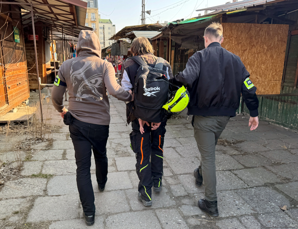 Trzech mężczyzn idzie wzdłuż pustego targowiska, jeden z nich jest prowadzony przez dwóch w mundurach.