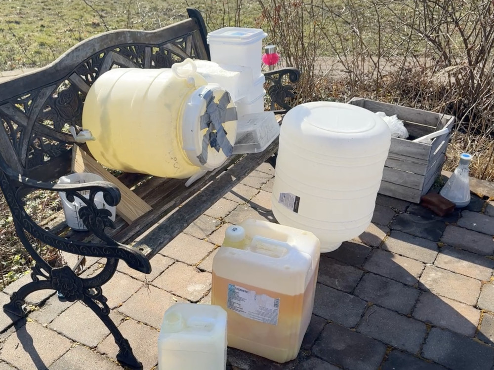 Na ławce w ogrodzie znajdują się plastikowe pojemniki, w tym żółty zbiornik z taśmą, obok drewniana skrzynka.
