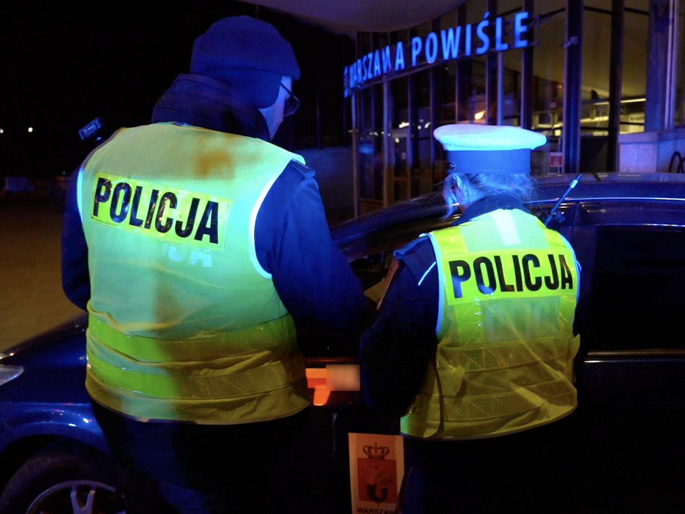 Policjanci w odblaskowych kamizelkach kontrolują pojazd nocą przed budynkiem stacji kolejowej.