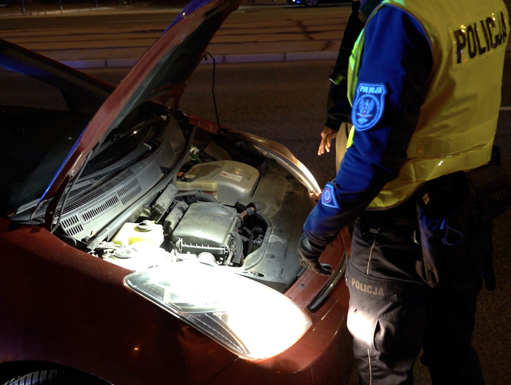 Policjant sprawdzający silnik czerwonego samochodu osobowego na drodze w nocy.
