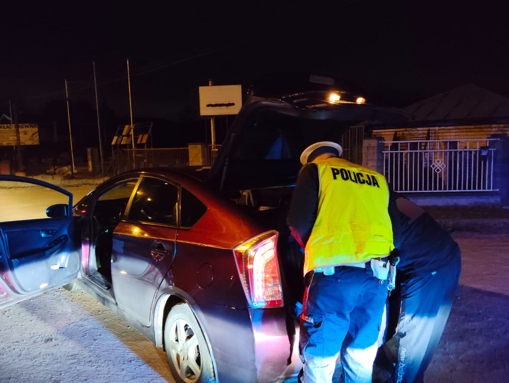 Policjant w odblaskowej kamizelce kontroluje bagażnik czerwonego samochodu osobowego w nocy.