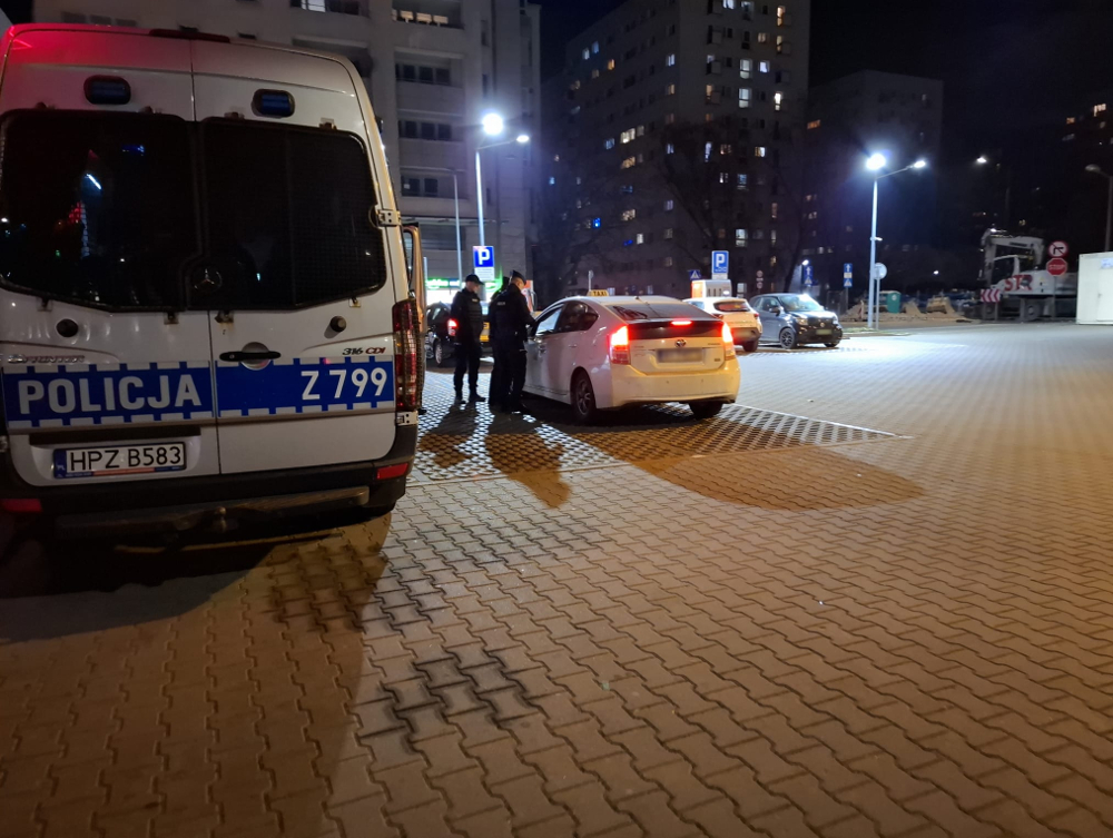 Policjanci rozmawiają z kierowcą białego samochodu osobowego na oświetlonym parkingu nocą.
