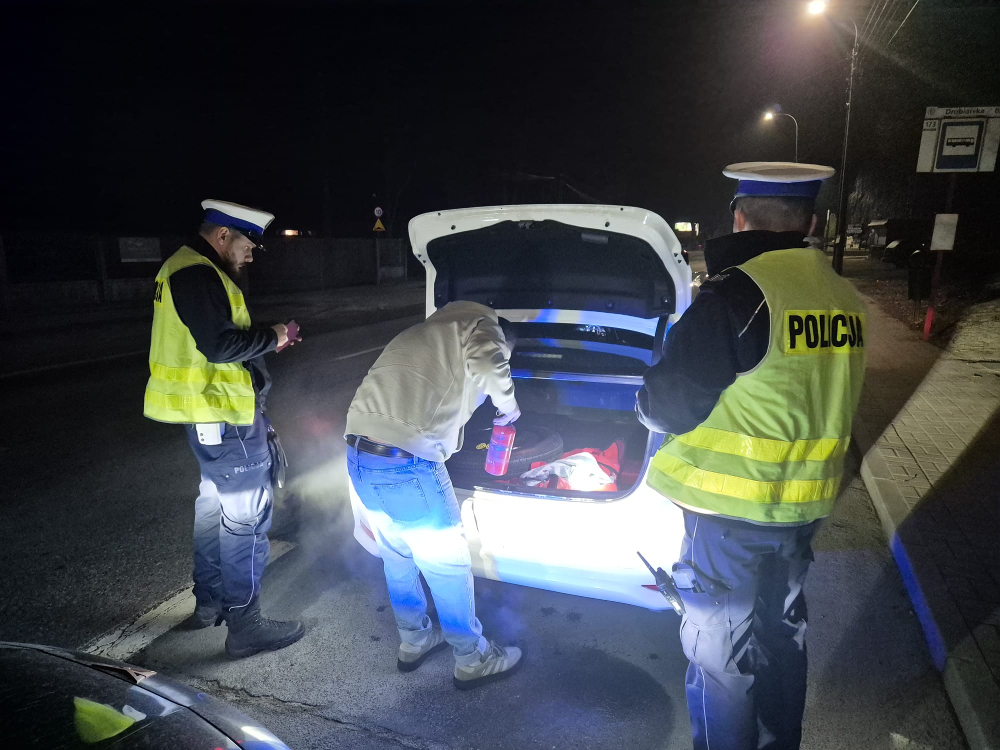 Mężczyzna przeszukuje bagażnik samochodu pod oświetleniem policyjnym, obok dwóch policjantów w kamizelkach odblaskowych.