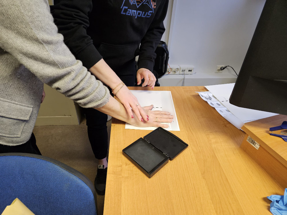 Dwie osoby wykonują odcisk palca na kartce papieru w biurze, obok znajduje się czarna poduszka do tuszu.