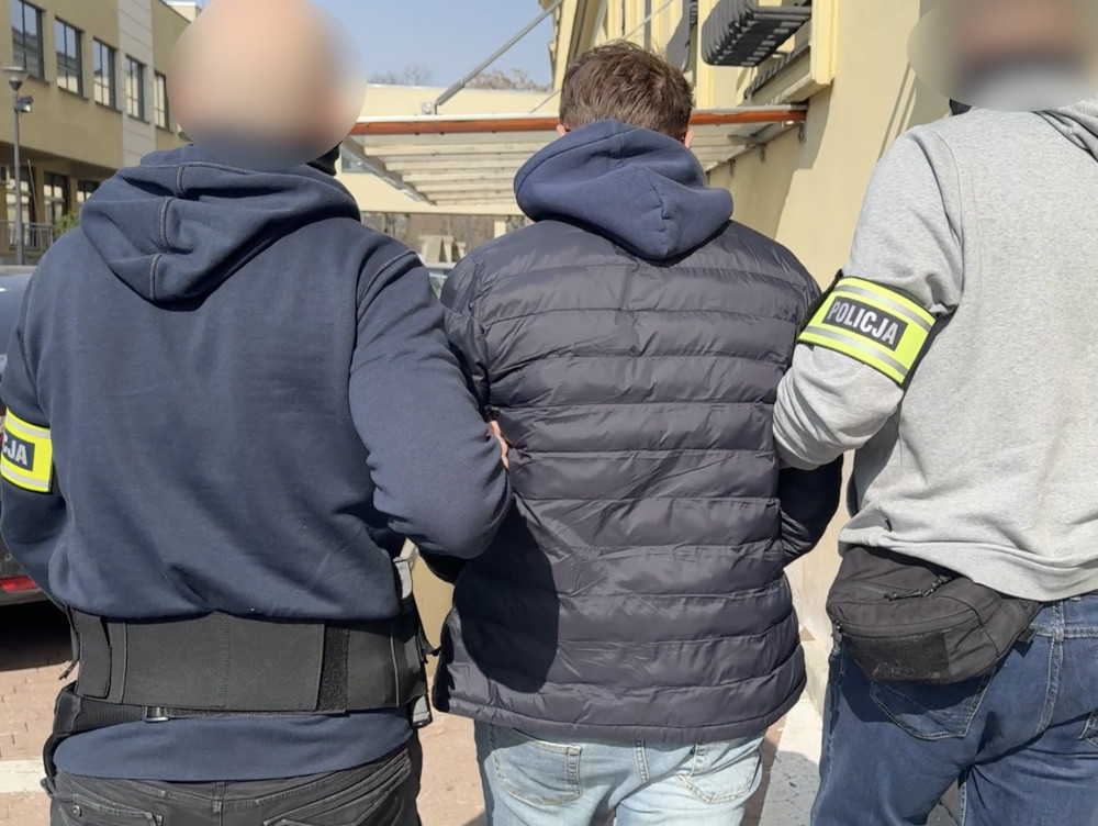 Mężczyzna w kurtce puchowej prowadzony przez dwóch funkcjonariuszy policji na terenie miejskim.