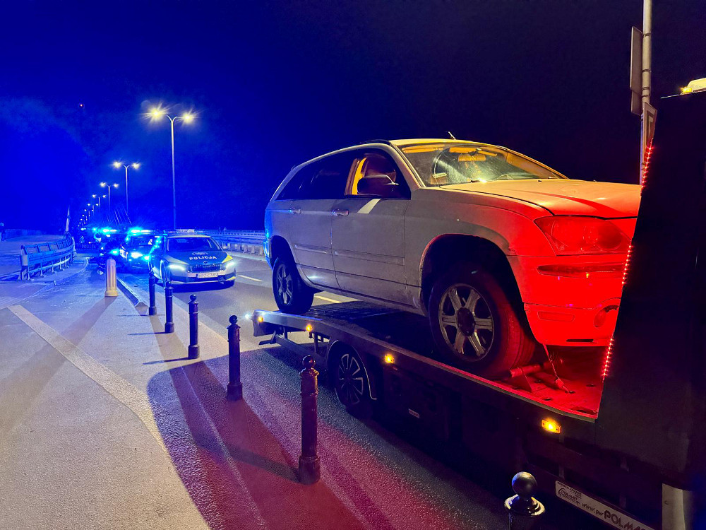 Biały SUV załadowany na lawetę, otoczony przez radiowozy policyjne z niebieskimi światłami na nocnej drodze.