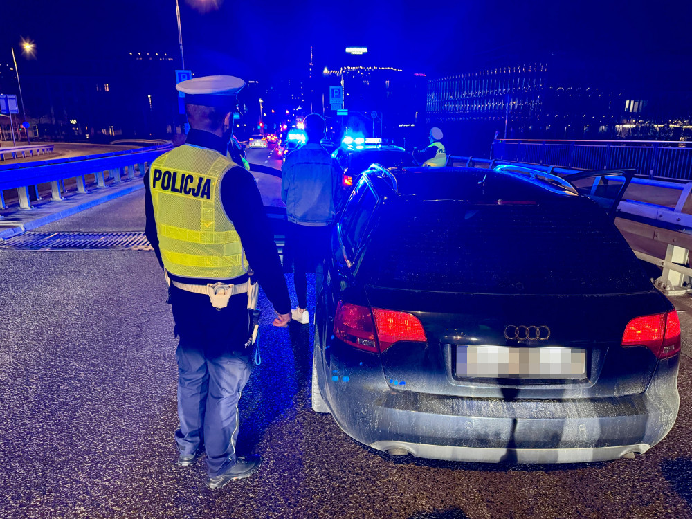 Policjant w kamizelce odblaskowej kontroluje kierowcę przy zatrzymanym samochodzie osobowym w nocy.