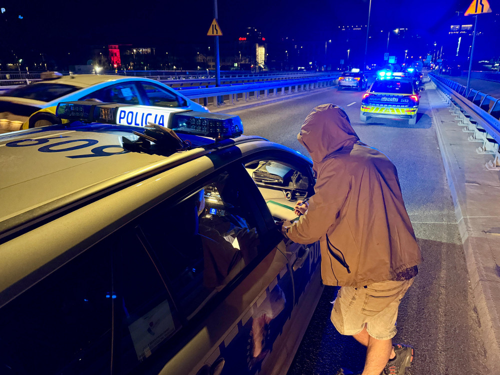 Mężczyzna w kapturze rozmawia z policjantem przy radiowozie na drodze w nocy, otoczeni przez inne pojazdy.