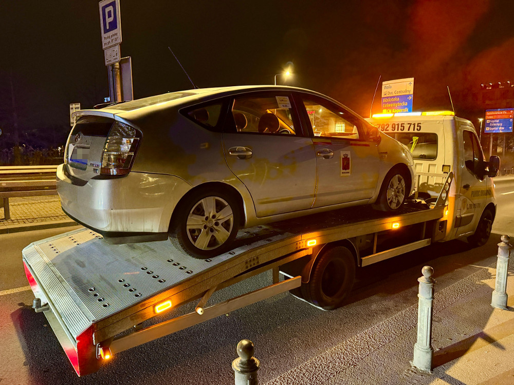 Samochód osobowy na lawecie, zaparkowany na poboczu drogi przy znakach drogowych w nocy.