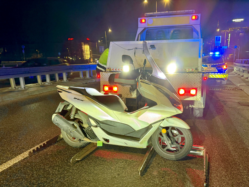 Skuter zaparkowany na drodze nocą, obok pojazdu holowniczego z włączonymi światłami.