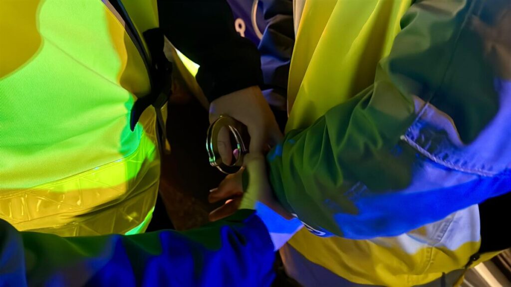 Funkcjonariusze policji zakładają kajdanki mężczyźnie w odblaskowej kamizelce w nocy.