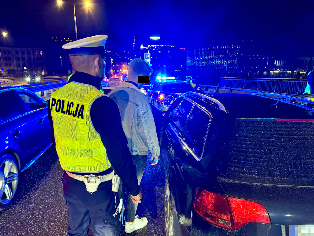 Policjant w kamizelce odblaskowej kontroluje mężczyznę na zatłoczonej drodze nocą, w tle widać radiowozy z włączonymi sygnałami.