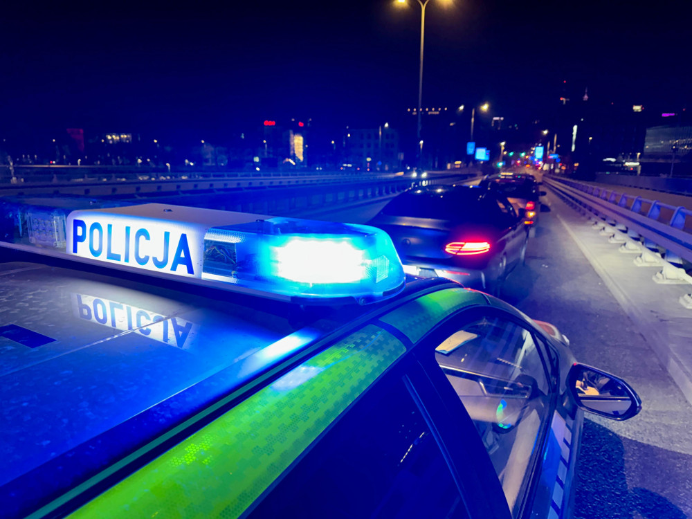 Policyjny radiowóz z niebieskimi światłami stoi na moście, a w tle widać kolejkę samochodów w nocy.