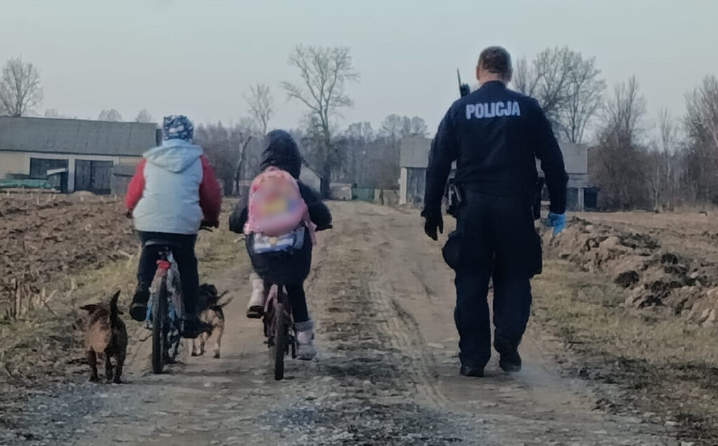 Dwoje dzieci na rowerach jedzie w stronę policjanta idącego po wiejskiej drodze. Obok towarzyszą im dwa psy.