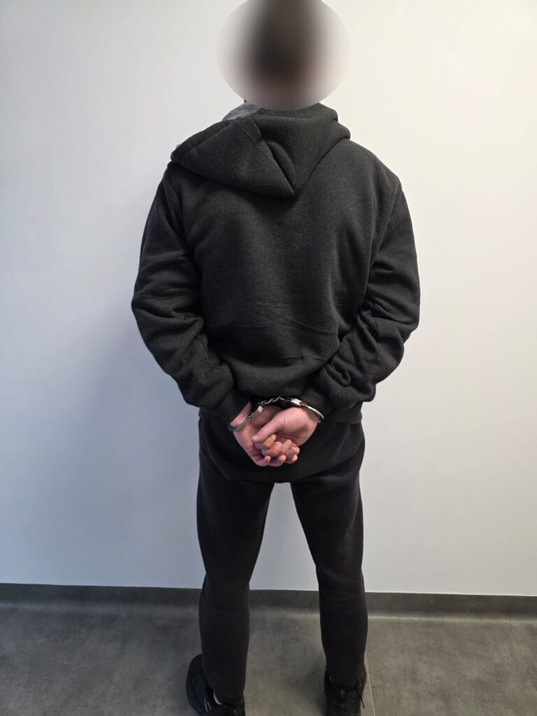 Mężczyzna w ciemnym dresie z rękami skuty w kajdankach stoi tyłem na tle jednolitej ściany.