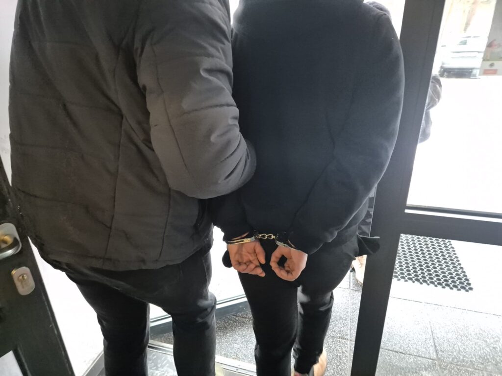 Mężczyzna w kajdankach prowadzony przez policjanta w drzwiach budynku.