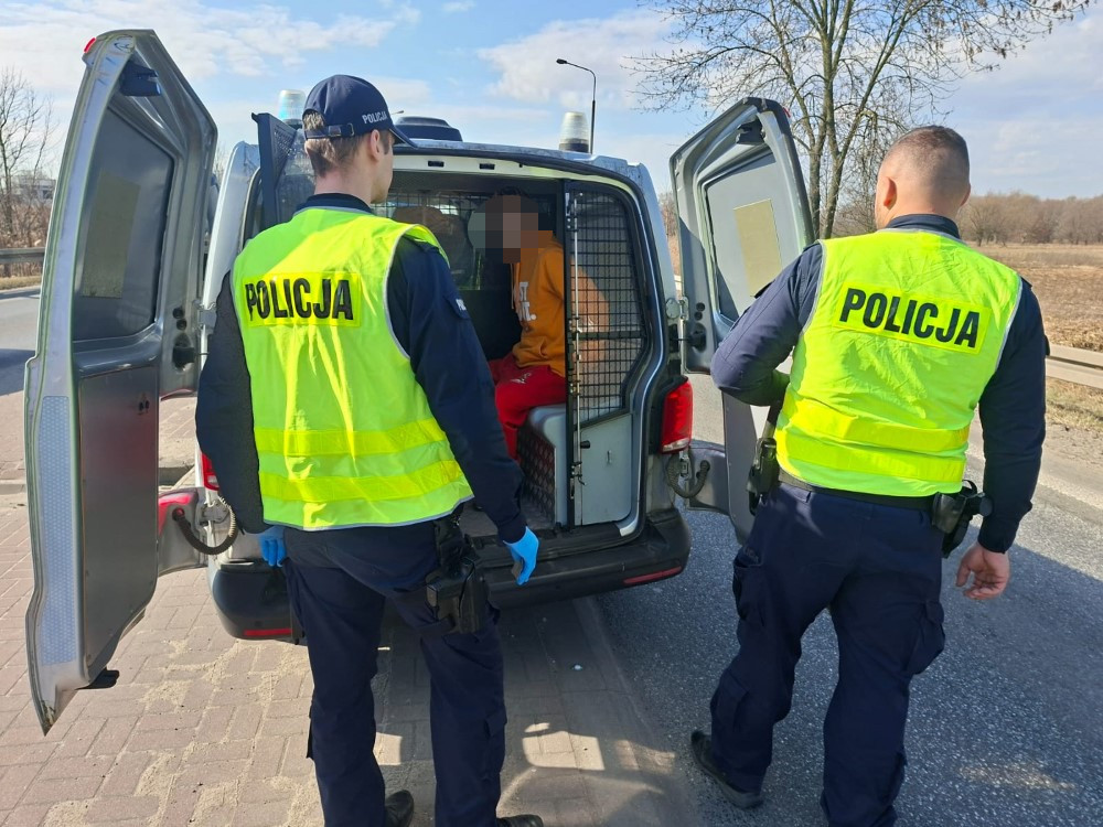 Dwóch policjantów w kamizelkach odblaskowych rozmawia z osobą siedzącą w furgonetce policyjnej na poboczu drogi.