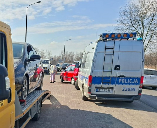Na drodze stoi żółta laweta z czarnym samochodem, obok zatrzymany radiowóz policyjny, a w tle widać korki.