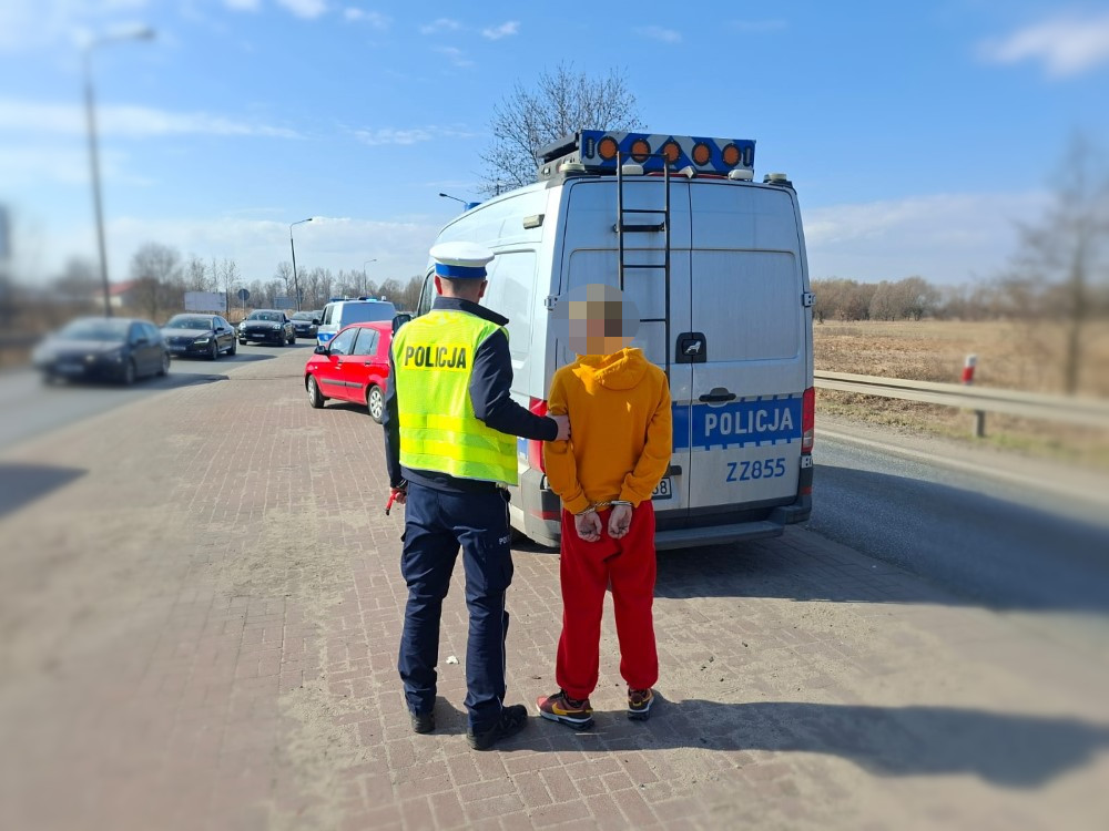 Policjant w kamizelce odblaskowej zatrzymuje mężczyznę w żółtej bluzie i czerwonych spodniach przy radiowozie.
