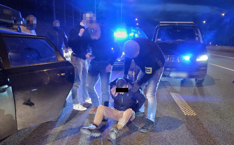 Grupa funkcjonariuszy w mundurach zatrzymuje mężczyznę siedzącego na jezdni w nocy, przy radiowozach z włączonymi sygnałami.