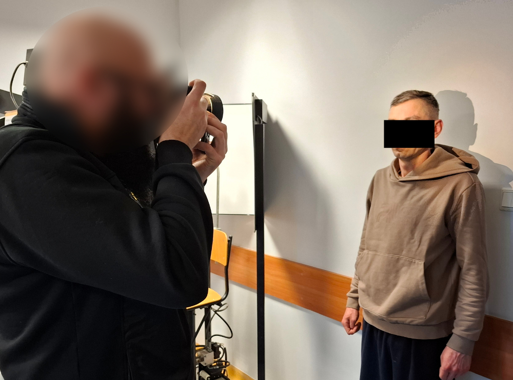 Mężczyzna w szarym dresie stoi w pomieszczeniu, podczas gdy inny mężczyzna z aparatem fotograficznym robi mu zdjęcie.