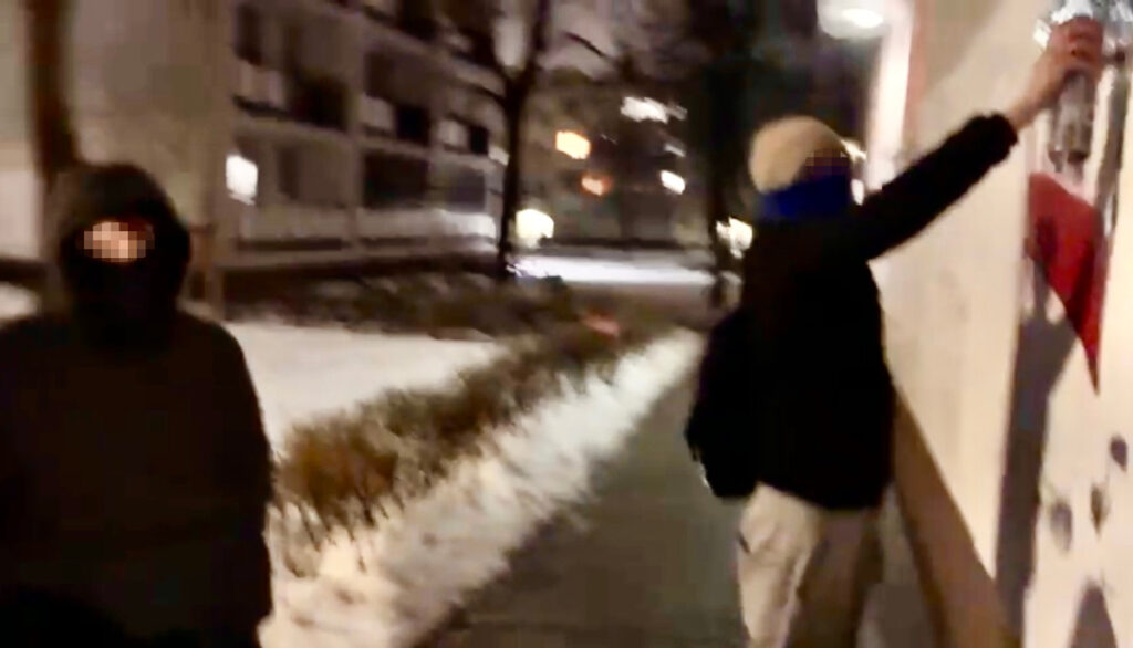 Mężczyzna w czarnej bluzie stoi na chodniku, podczas gdy inny mężczyzna w jasnej czapce maluje na ścianie budynku.