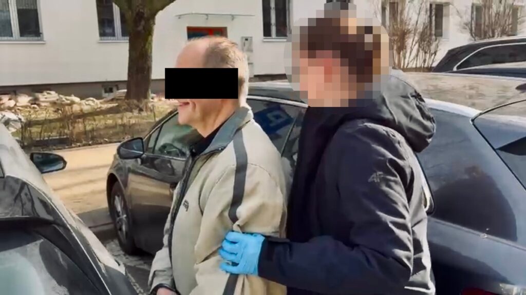 Mężczyzna w szarym dresie stoi obok samochodu, trzymany przez osobę w granatowej kurtce i rękawiczkach.