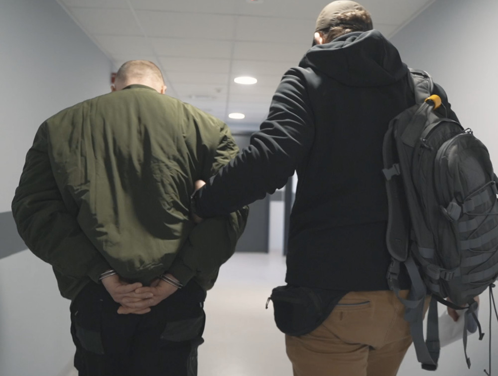 Mężczyzna w kajdankach prowadzony przez funkcjonariusza w korytarzu budynku.