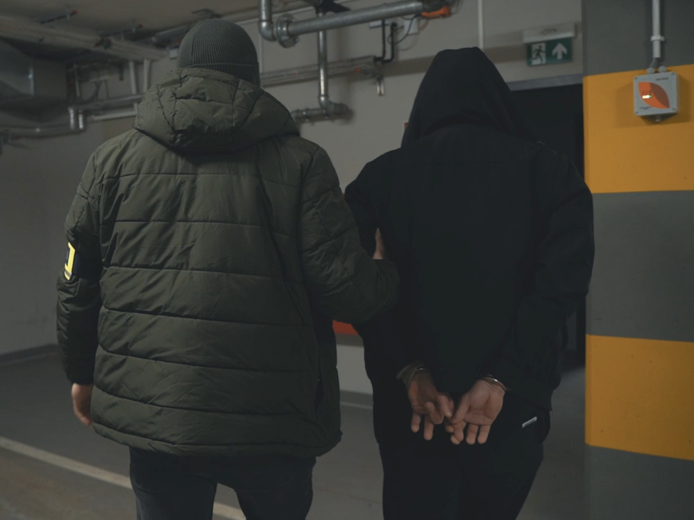 Funkcjonariusz w zielonej kurtce prowadzi zatrzymanego w kajdankach w podziemnym parkingu.