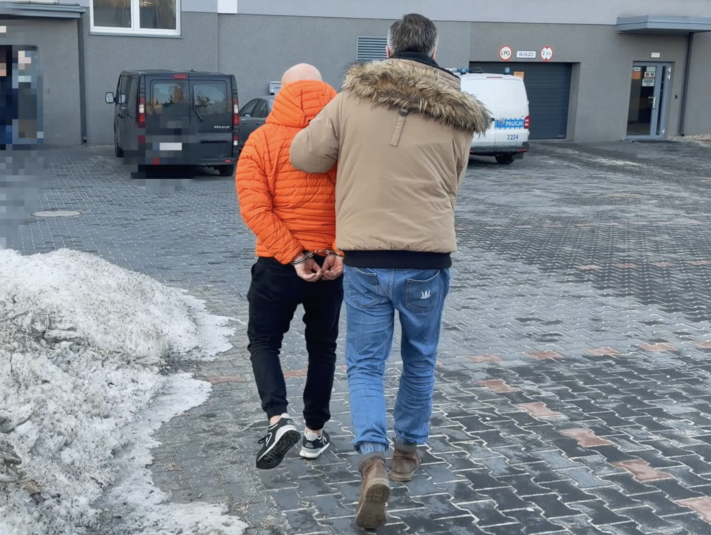 Mężczyzna w pomarańczowej kurtce, w kajdankach, prowadzony przez funkcjonariusza policji na parkingu.