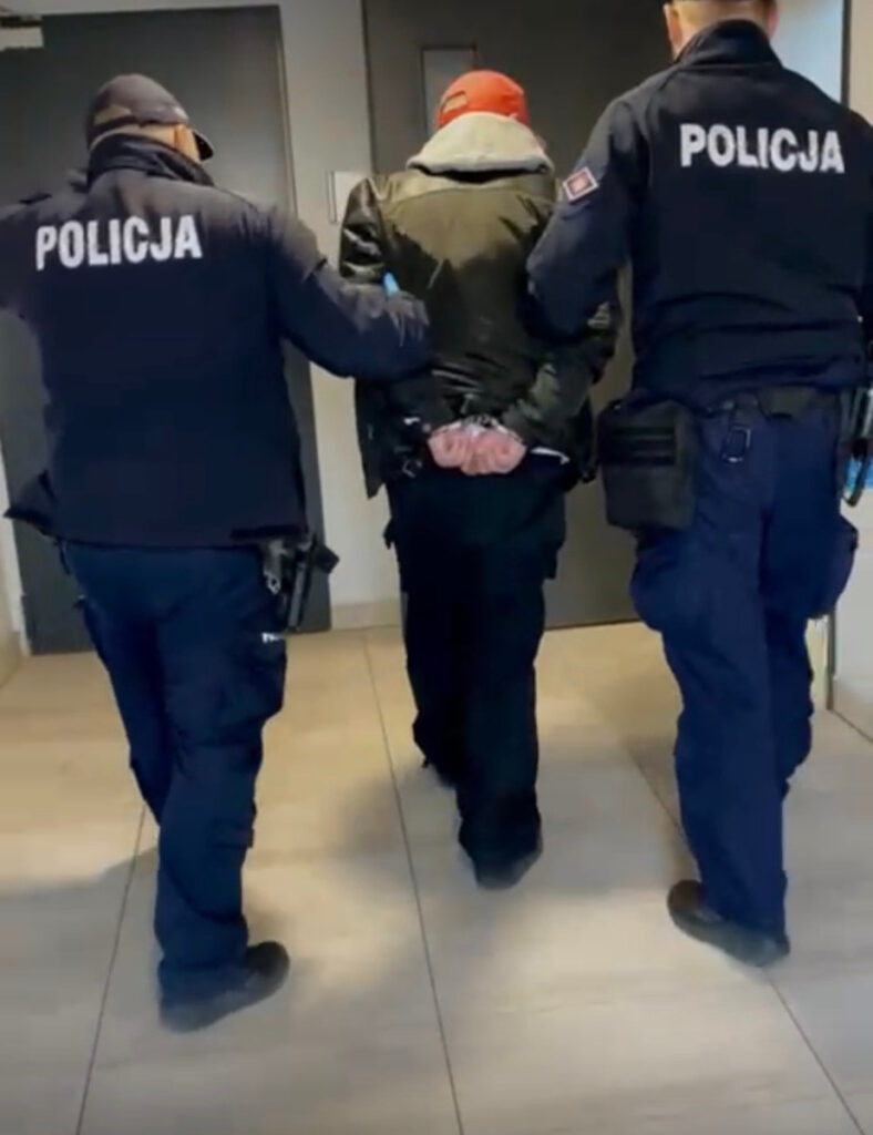 Mężczyzna z rękami związanymi z tyłu prowadzony przez dwóch policjantów w korytarzu budynku.