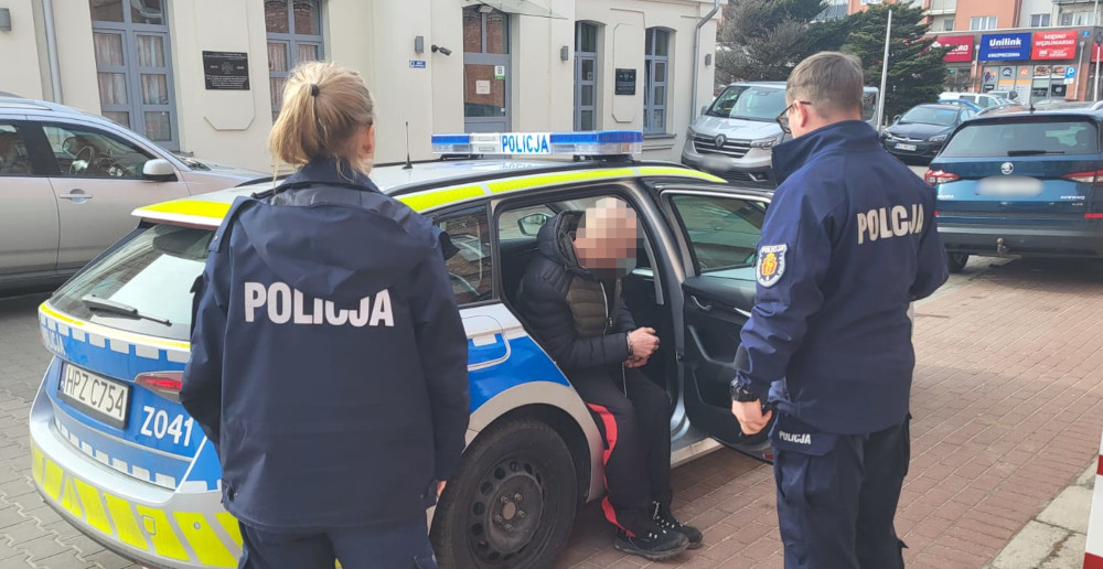 Policjantka rozmawia z mężczyzną siedzącym w radiowozie, na tle miejskiego krajobrazu.